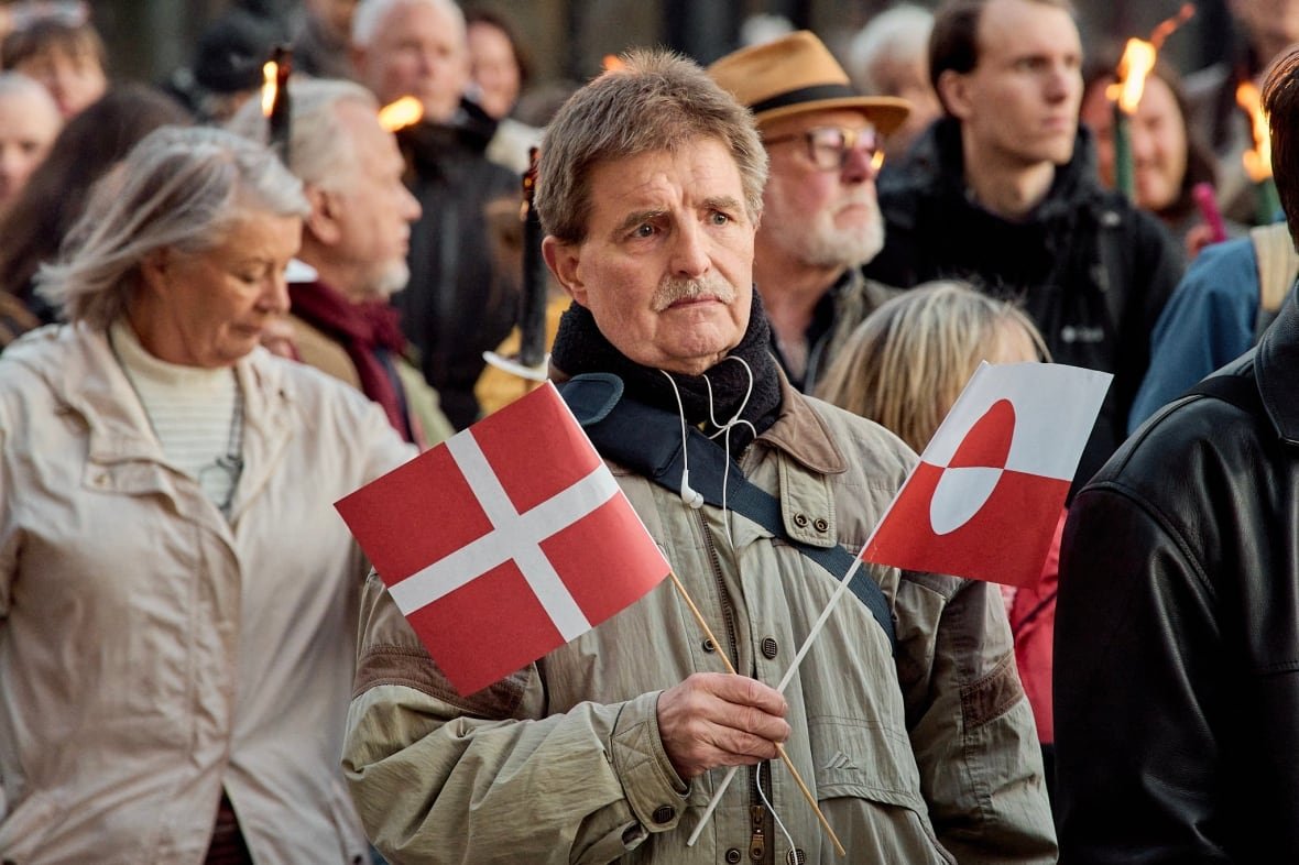 man-holding-a-denmark-flag-and-a-greenland-flag-at-demonstration-in-support-of-greenland A man holds a Denmark flag and a Greenland flag, while attending a demonstration in support of Greenland in Copenhagen.