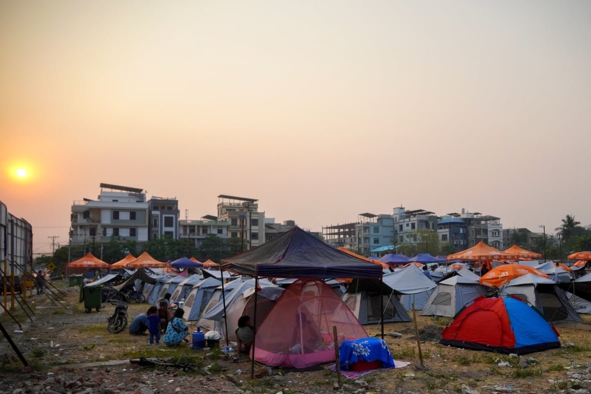 myanmar-quake-3 Rows of tents in an open field with damaged buildings in the background