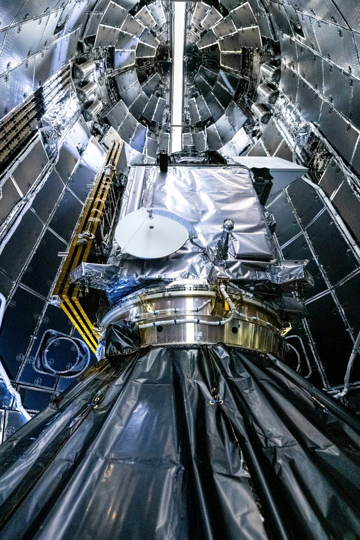 nasa-satellite-launch A satellite dish sits on an cylindrical base, viewed from inside a larger cylinder with many rectangular panels.