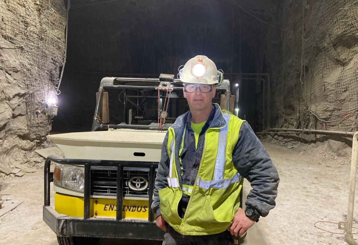nutrien-mine-employee-harry-booth-works-underground Miner dressed in safety gear wearing hard hat with head lamp is standing in front of Toyota truck inside one of the tunnels in the Picadilly mine.