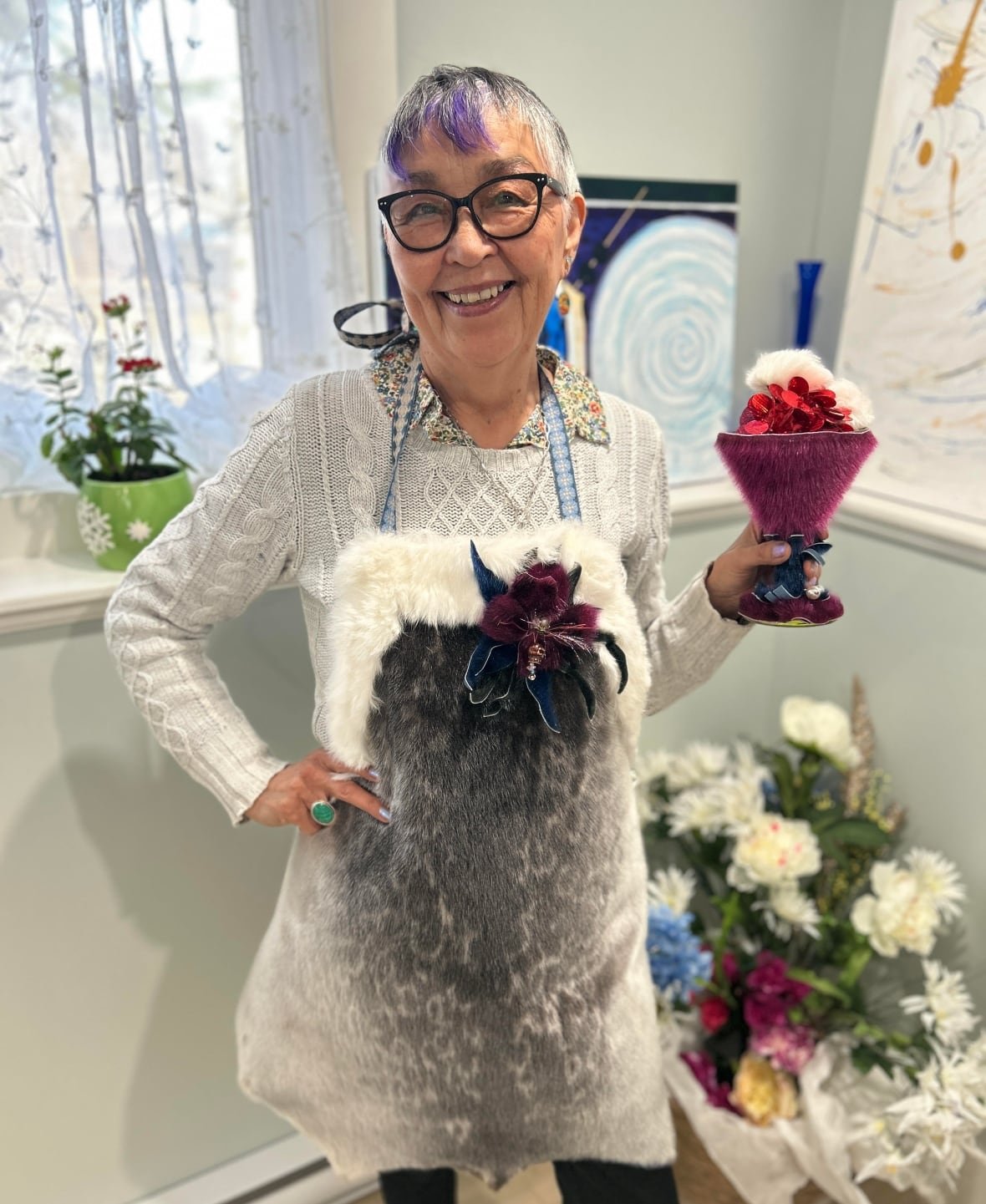 shirley-ensemble An Inuk woman in her 60s with short grey and purple hair wears a sealskin apron and holds a magenta seal skin wine glass.