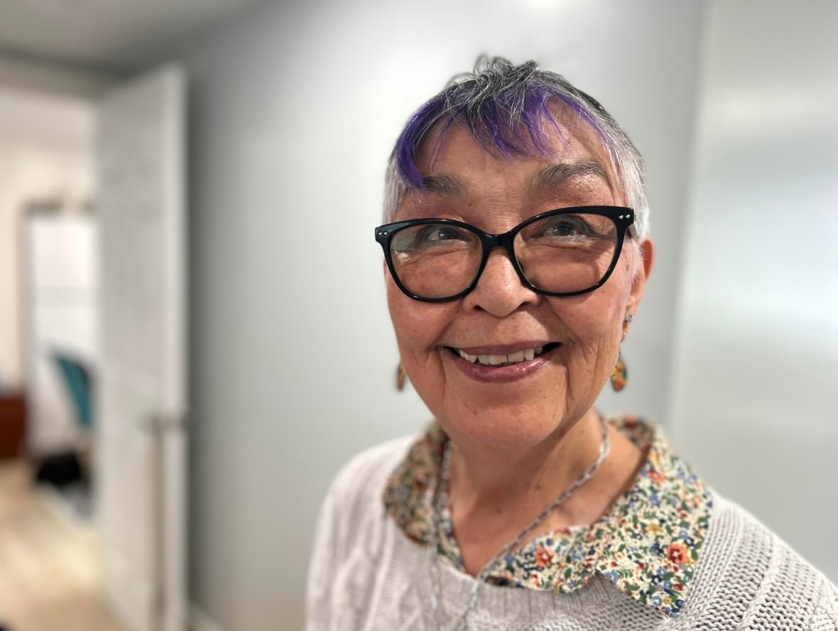shirley-moorhouse-profile-1 A woman in her 60s with grey and purple hair smiles for the camera. She is wearing a white sweater and a flowered collar.