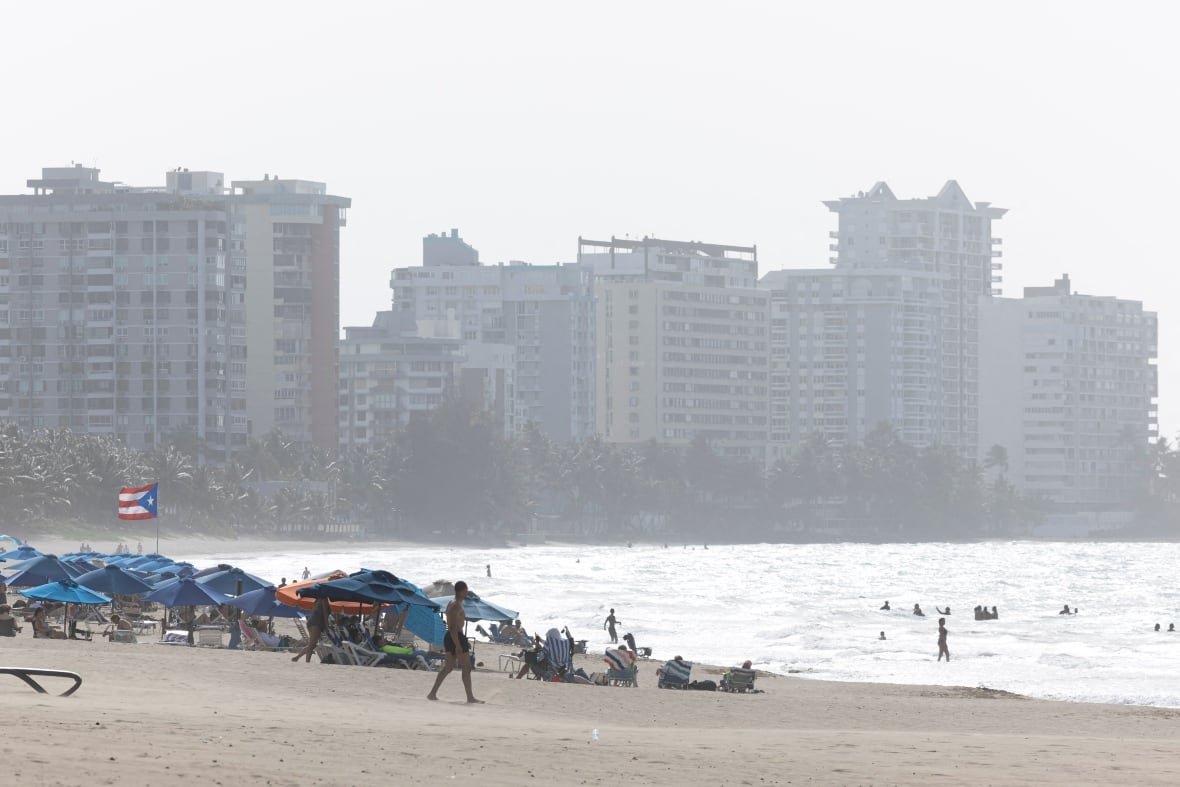 storm-dust-caribbean People some under beach umbrellas enjoy a beach as a cloud of dust from the Sahara Desert blankets Puerto Rico.