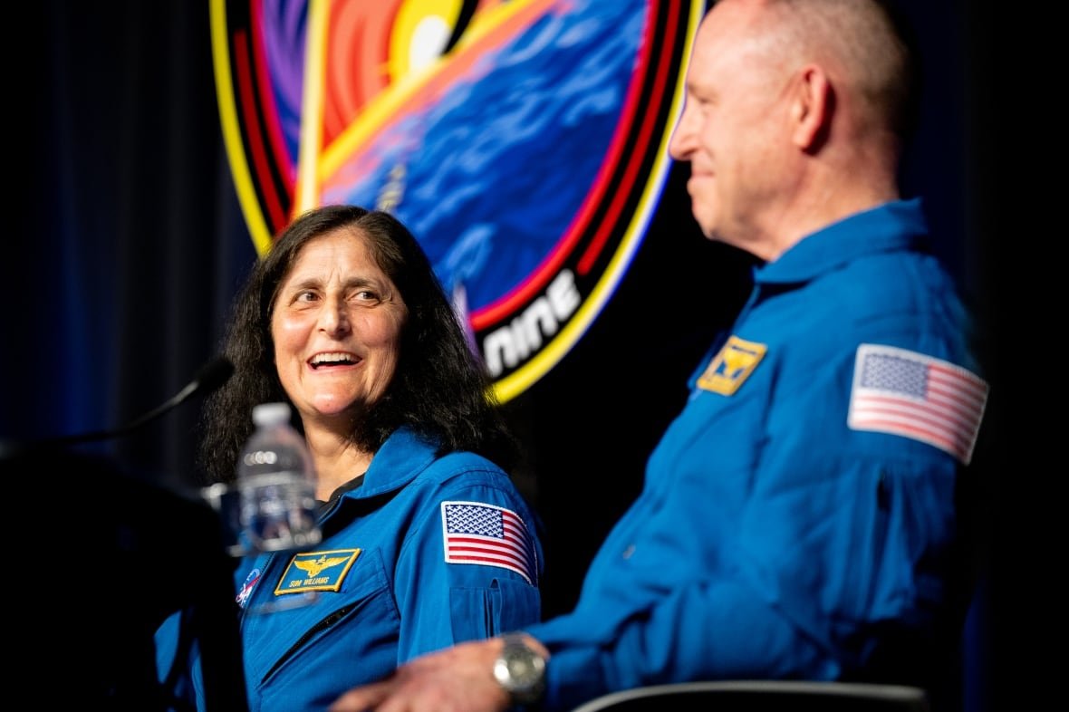 suni-williams-and-butch-wilmore-speak-1 Two people dressed in blue flight suits, with American flag patches on the arm, are shown seated on a stage.