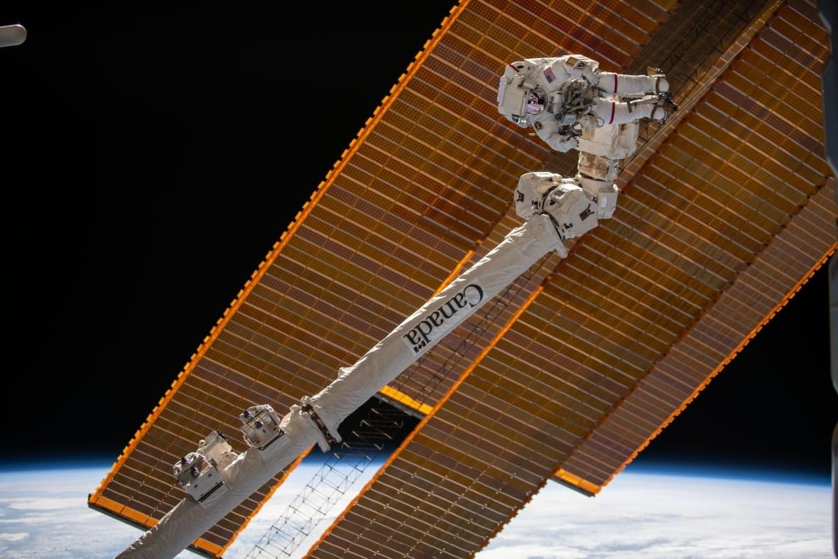suni-williams-spacewalk-canadarm An astronaut in a white spacesuit is attached to Canadarm 2 with the solar ray antennas in the background along with a sliver of Earth.