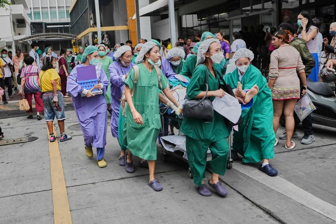 thailand-southeast-asia-earthquake Several people wearing hospital gowns and head coverings, mostly women, are shown outside a building, wheeling an unseen patient on a gurney.