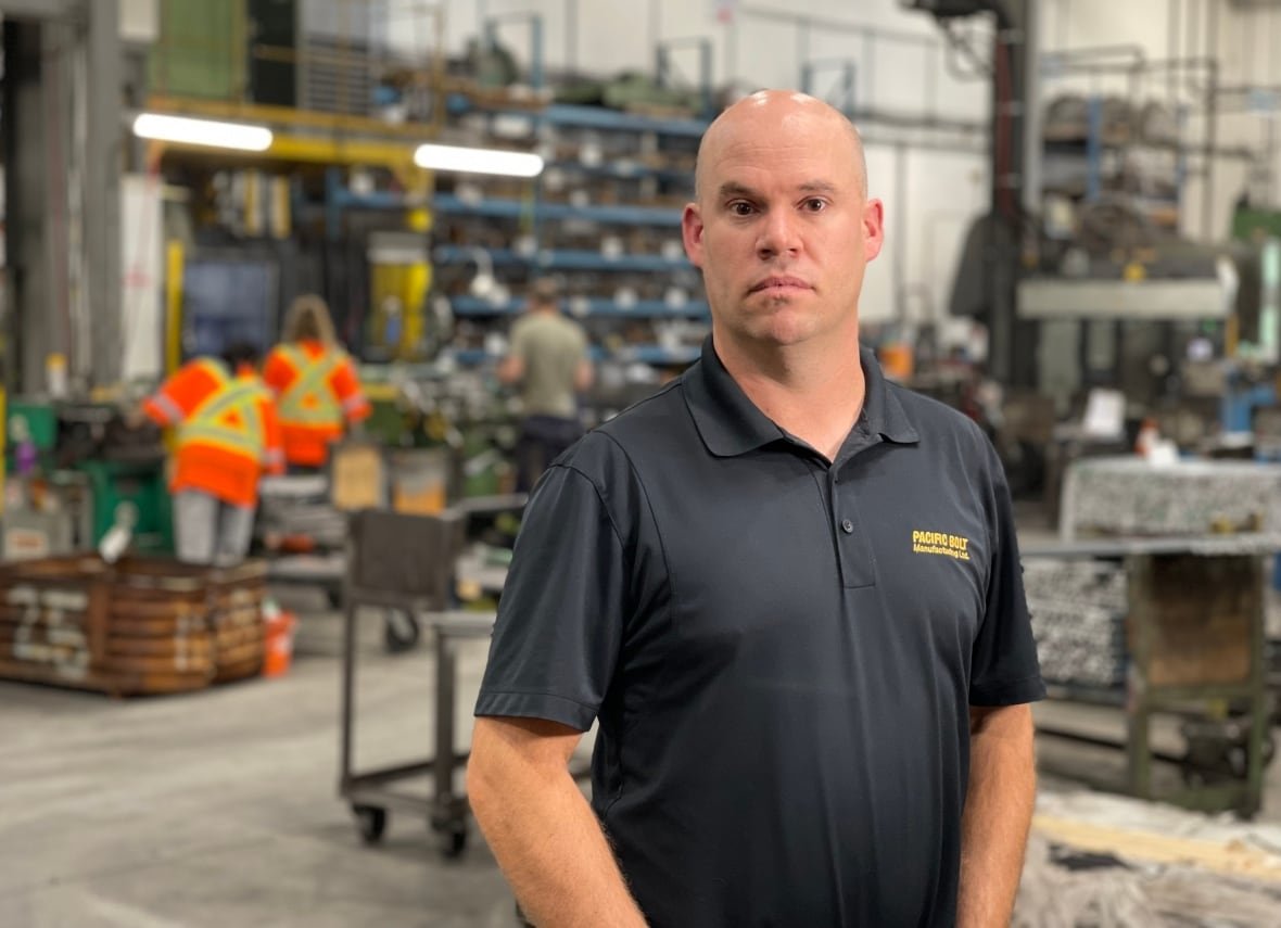 trevor-borland A bald man in a black polo poses for a photo in a factory floor setting as people in flourescent safety gear work behind him.