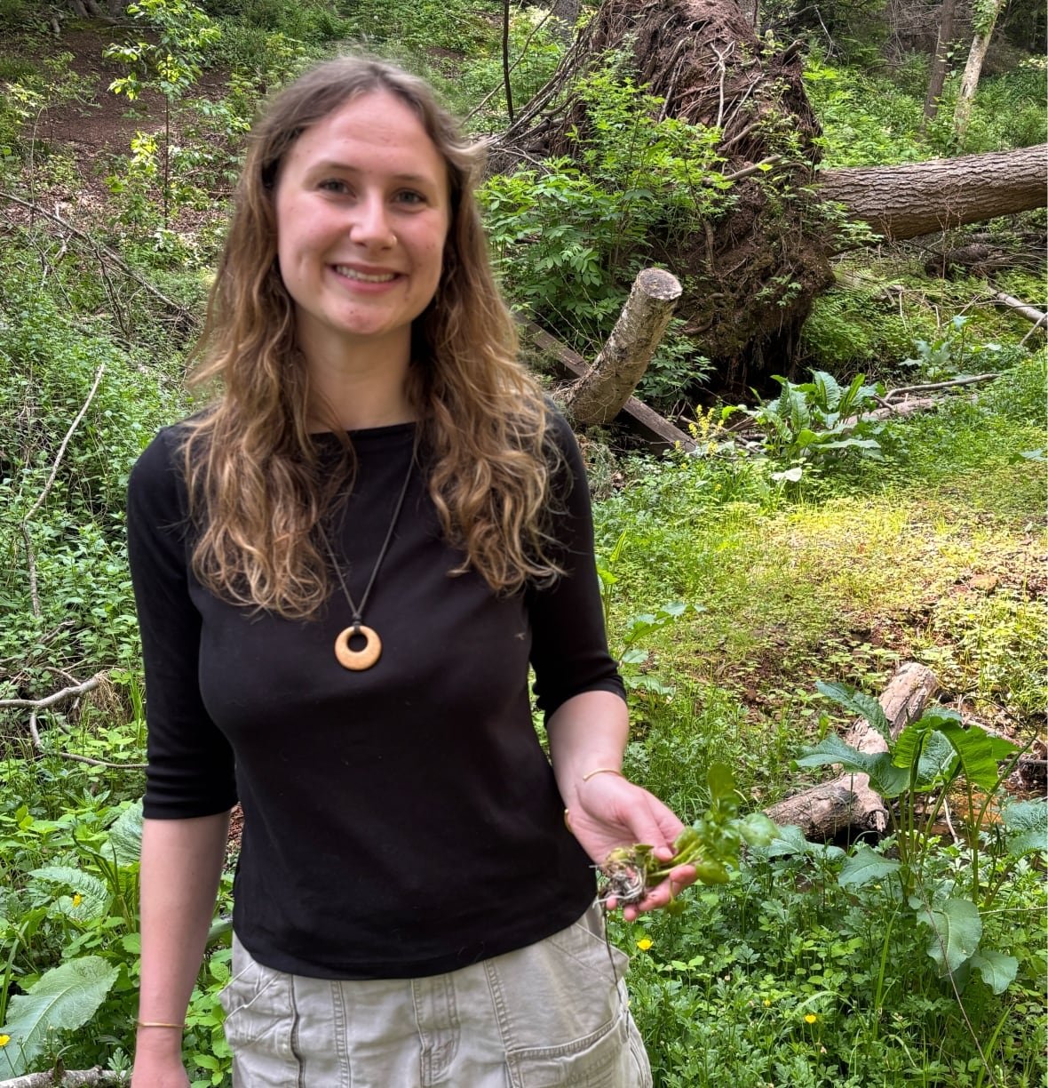 watercress-1 A woman stands outdoors in a green, wooded area, smiling and holding a small watercress plant in her hand. She is wearing a black top, light shorts, and a circular pendant necklace