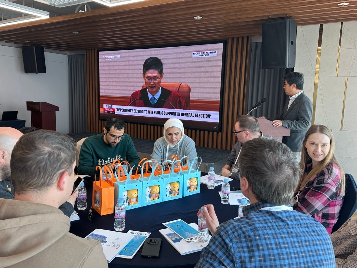 world-journalist-conference-south-korea A group of people sit around a table with a television in the background broadcasting a court ruling.