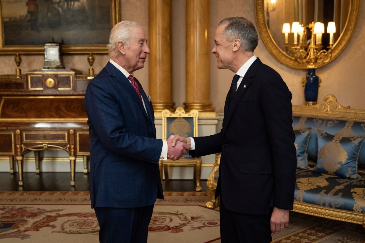 britain-canada-carney Two people shake hands standing in the centre of a room with furniture against the wall and lights reflected in a mirror.