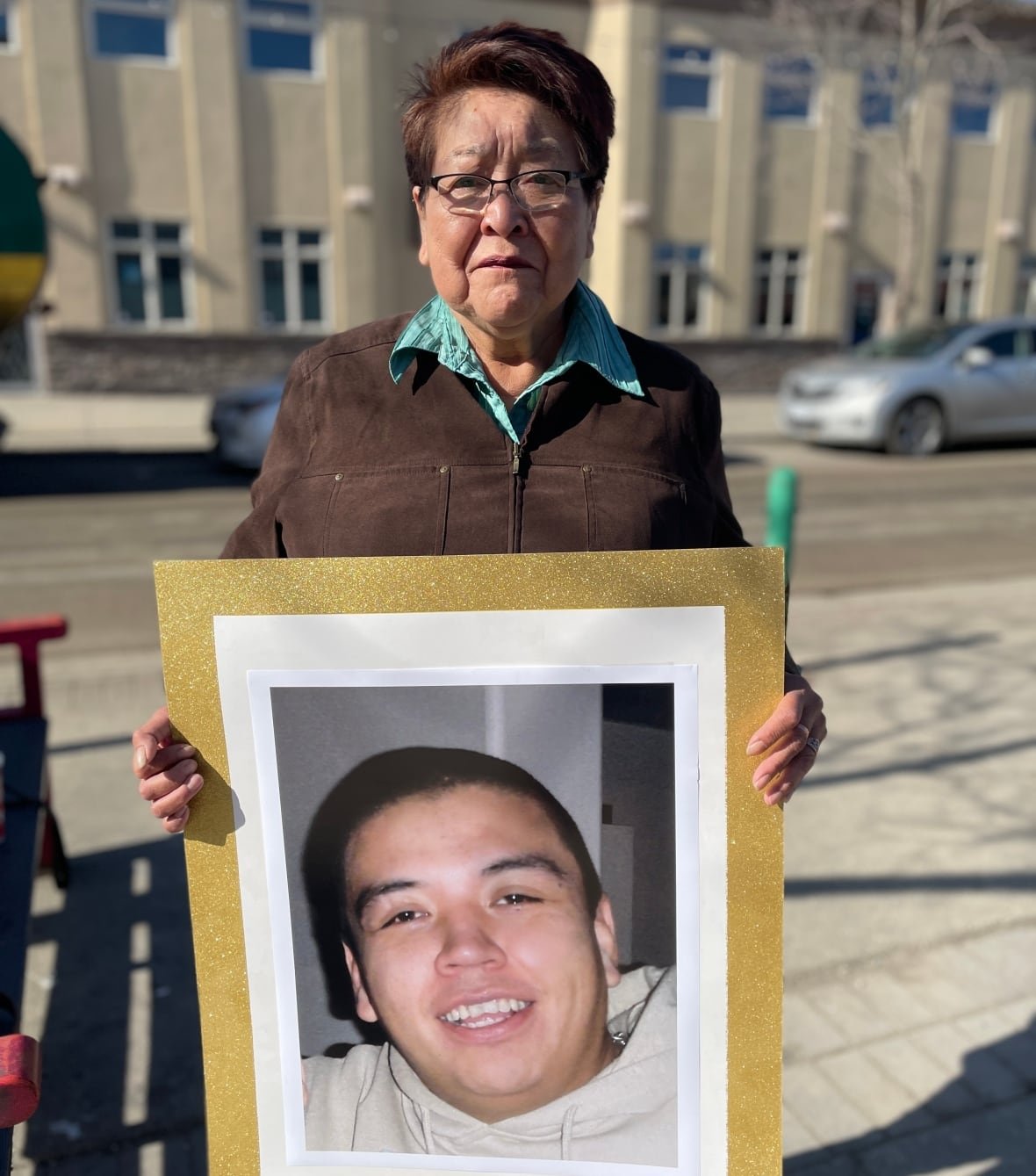 dale-culver-and-aunt An Indigenous woman holds a framed picture of a smiling man outside a building.
