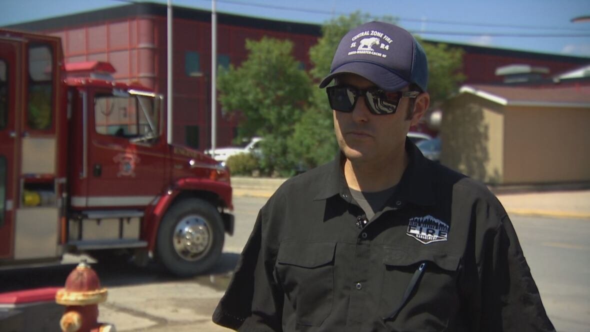 Man wearing navy blue hat and dark sunglasses wears a black button up short and stands in front of a red fire truck.