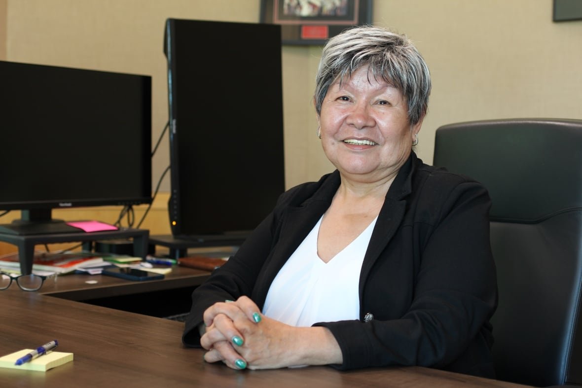 eva-clayton A woman in a black blazer and white blouse poses for a photo while seated at her desk.