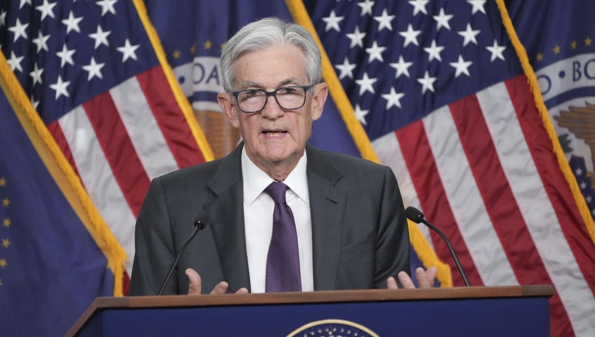 A man with grey hair stands at a podium in front of a bunch of American flags. He is in the middle of speaking and has his hands slightly raised.