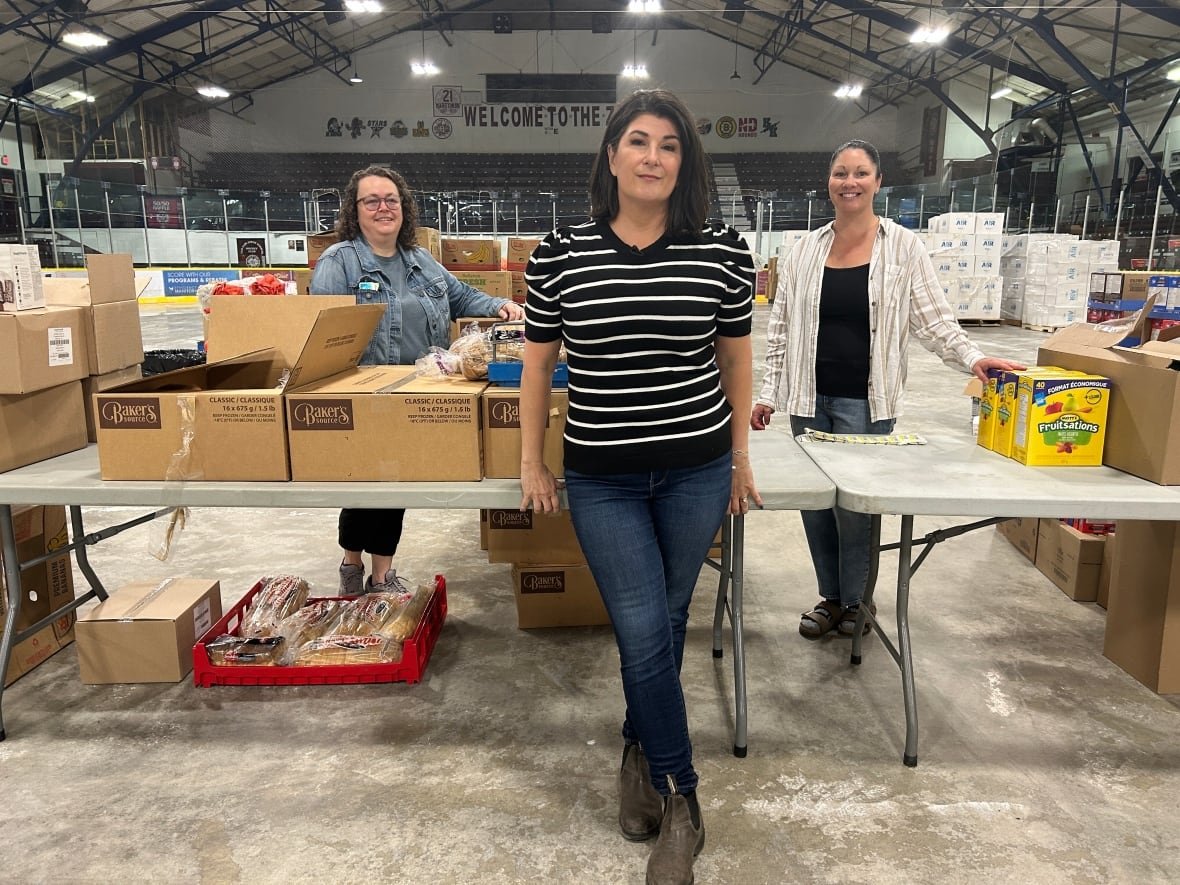 flin-flon-deputy-mayor-alison-dallas-funk-evacuees-return Three women stand next to tables covered with boxes of donated items in a hockey arena.