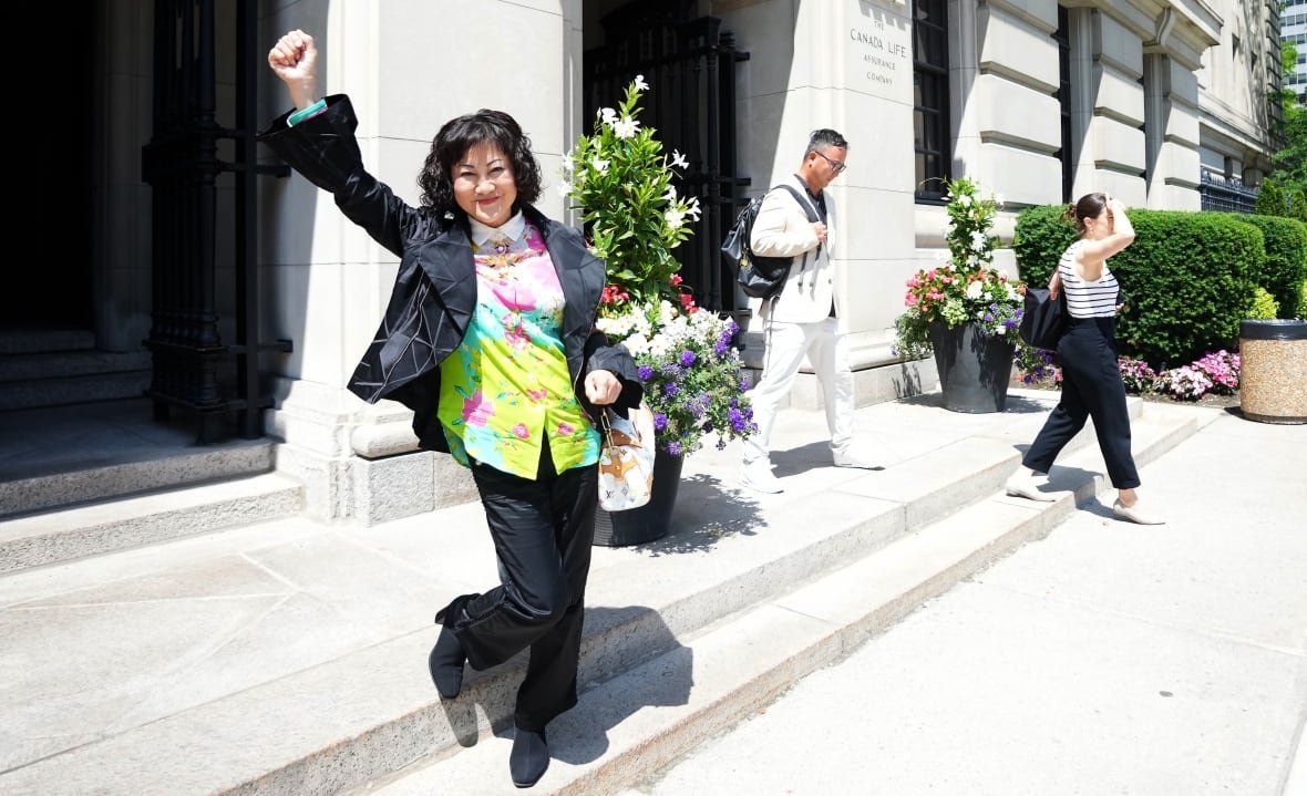 A woman with a black bob, black jacket, floral shirt and black pants pumps a fist in the air, standing on the steps of a courthouse.