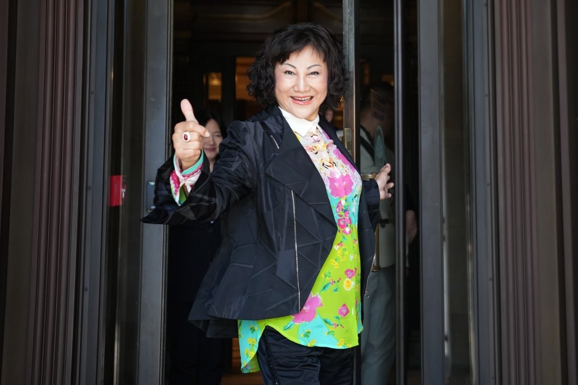A woman with black hair in a bob, wearing a black coat over a bright, floral top, gives a thumbs up sign.
