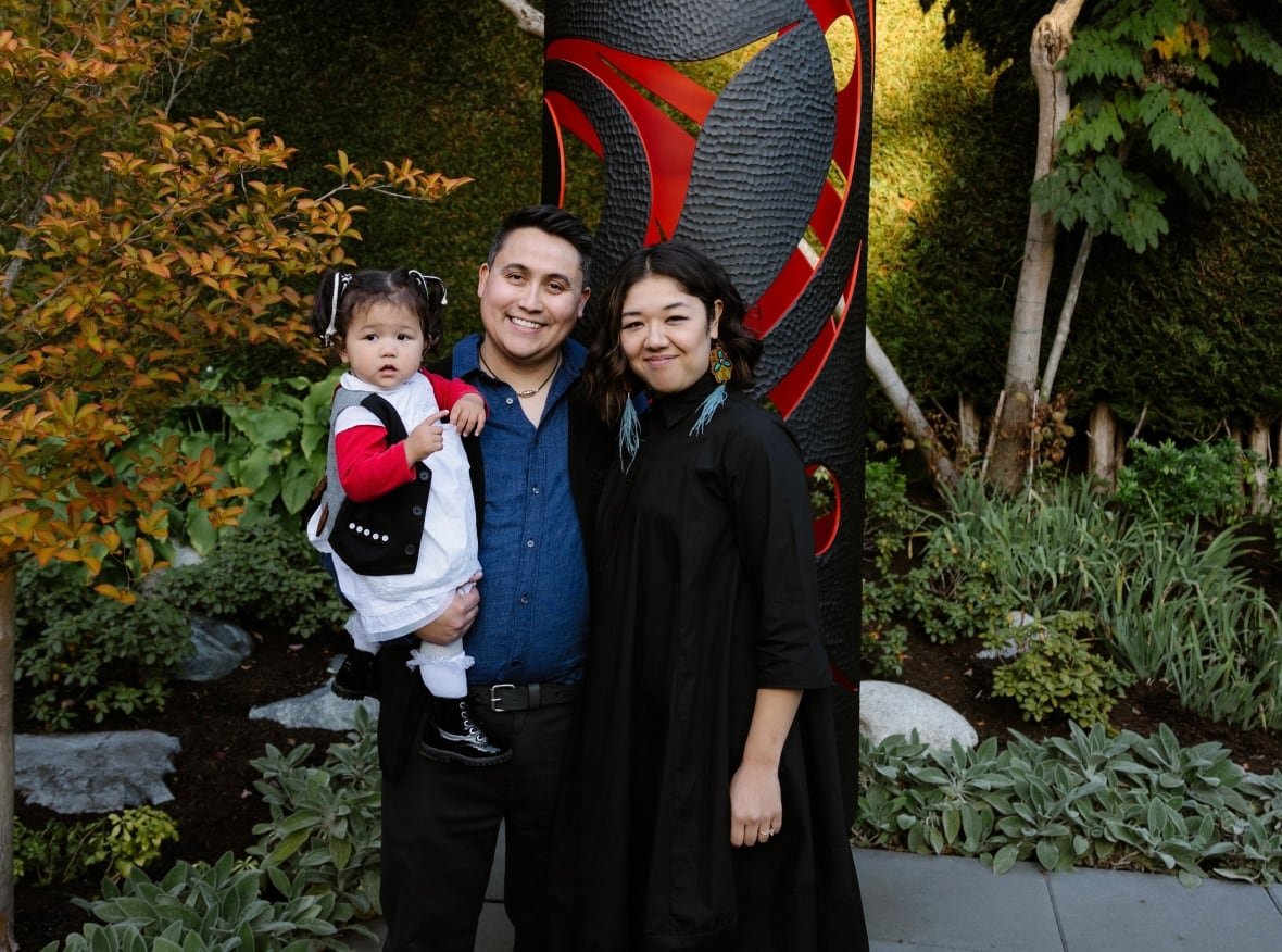 james-harry-lauren-brevner-and-their-daughter-hana A man and woman post for a photo holding their young daughter in front of an art piece.