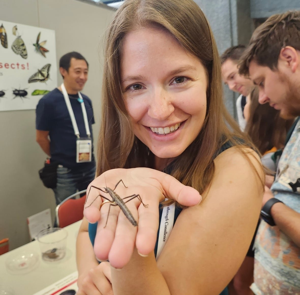 A woman smiling at the camera holding a bug, with people looking at a bug exhibit in the background.