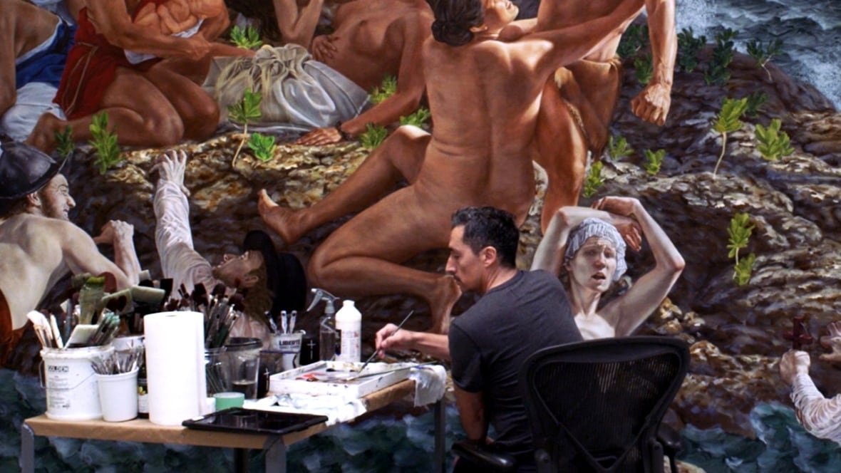A man at a table full of paint brushes and paint, painting a large mural featuring naked people on a rocky shore.