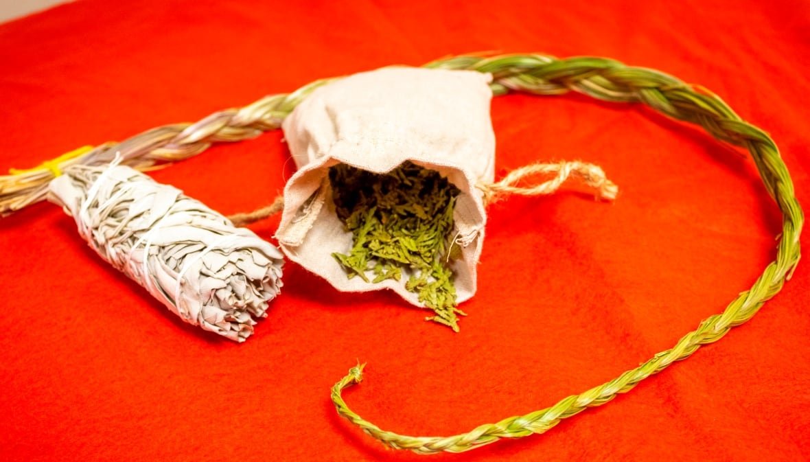 sacred-medicines Sacred Indigenous medicines, including sweetgrass and white sage, laid out on a red tabletop.
