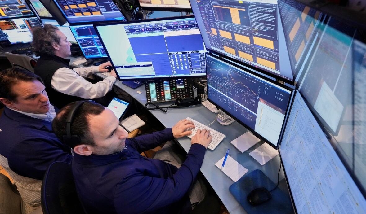 A partially overhead view looks down on traders sitting at a computer setup with a bunch of monitors above and around them, displaying various graphs. The traders look concentrated. There are three of them, two looking at the same setup, one looking at his own setup in the background.