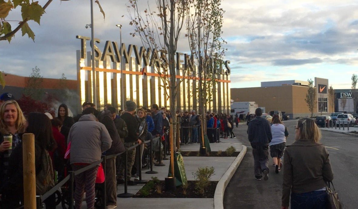 A lineup of people outside a large mall with a sign reading 'Tsawwassen Mills.'