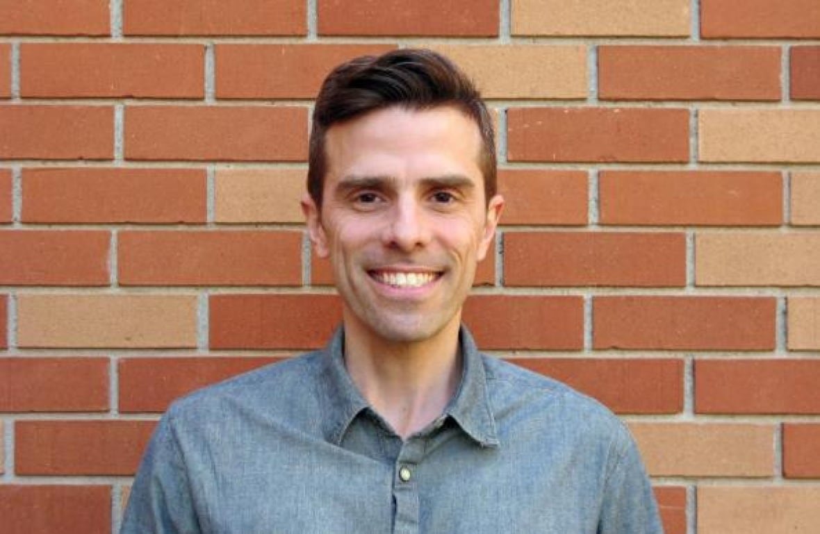 u-of-m-prof-ryan-cardwell A man with dark brown eyes and dark brown hair wearing a blue button-up shirt stands in front of a brick wall and smiles.