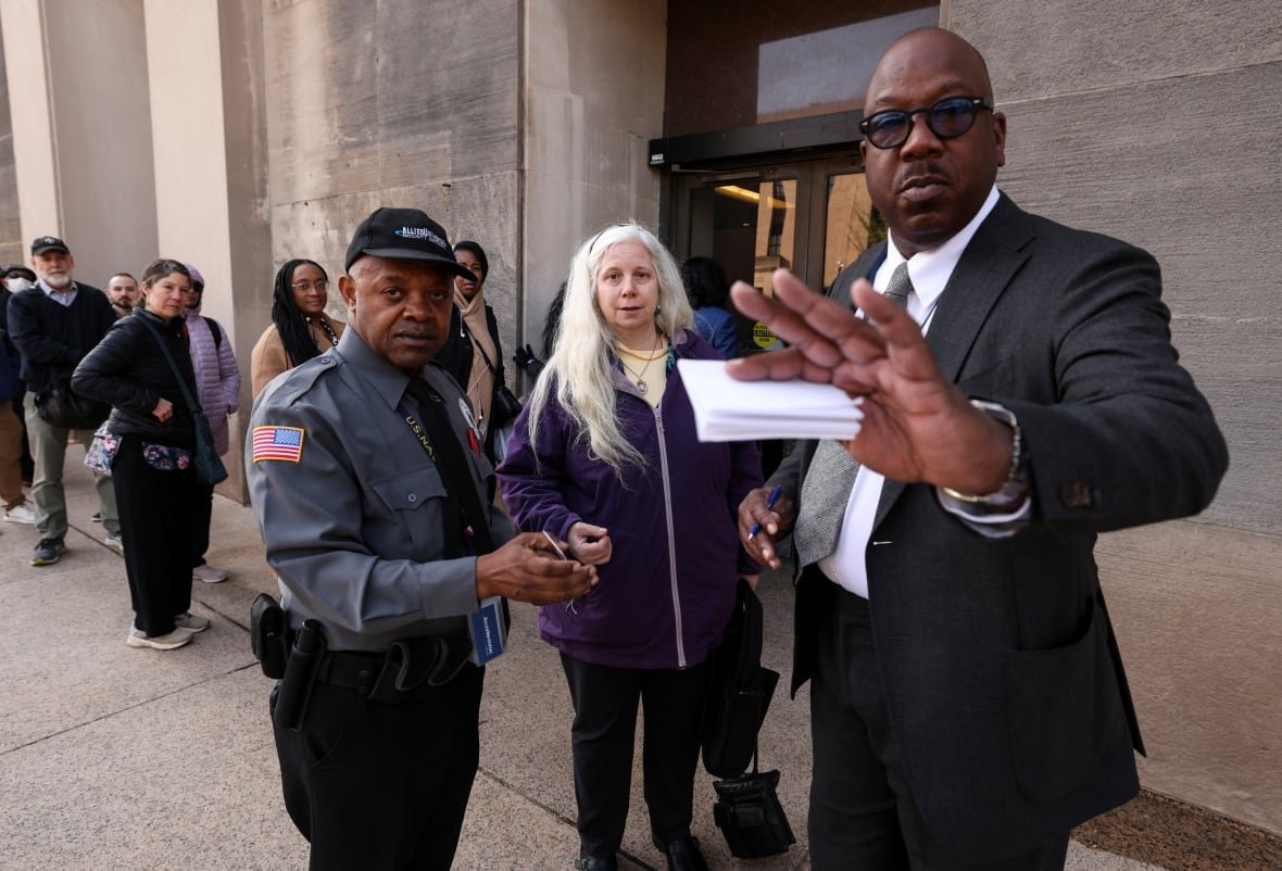 usa-trump-health A woman with white hair stands outside a government building looking stunned. Two security guards stand to her side, one gestures at the camera.