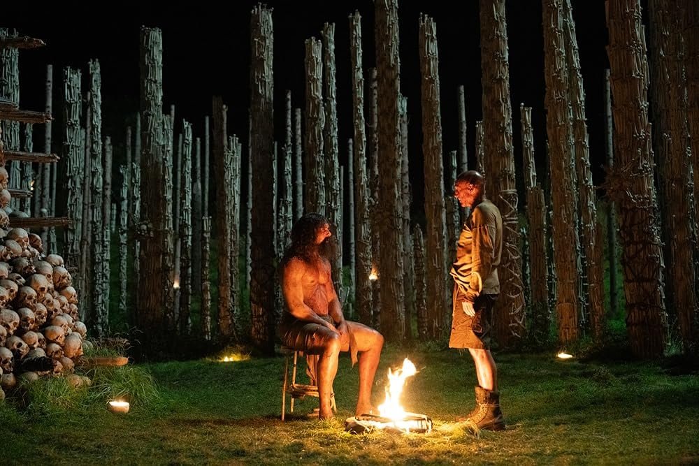 A seated, shirtless man is surrounded by a pile of bones and a large group of pillars. Next to him stands a man in dirty clothes.