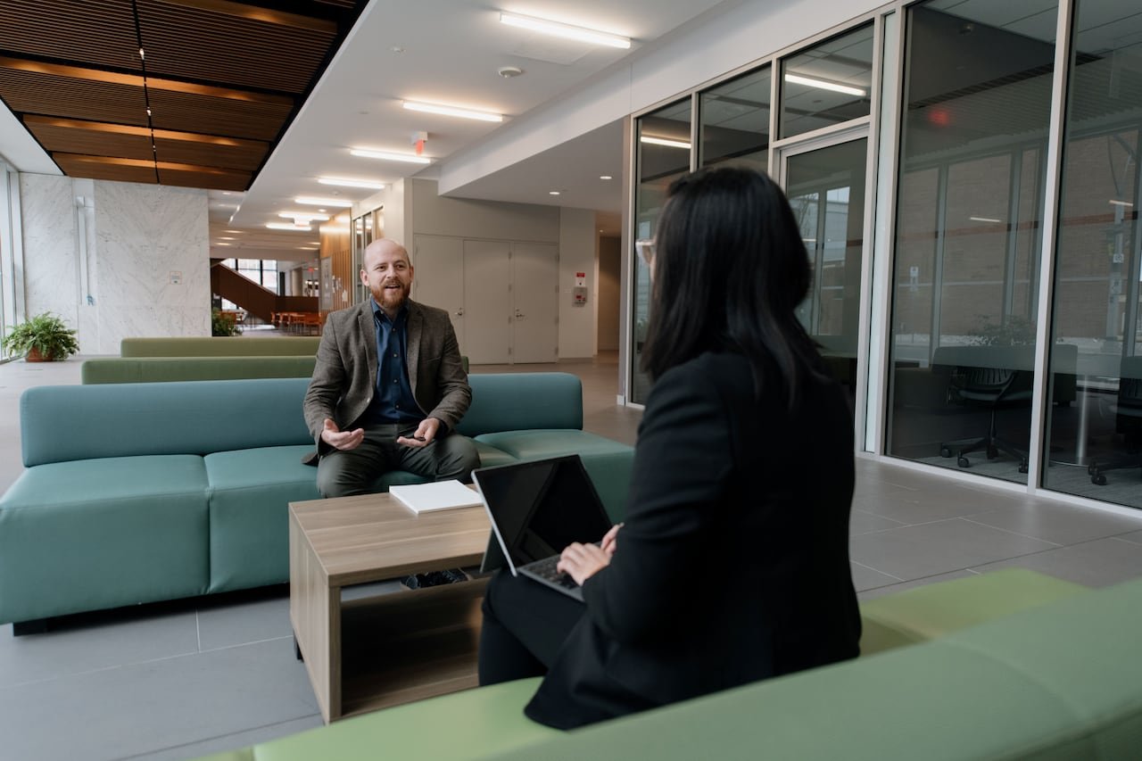 A bald man with a brown beard in a suit sits across from a young woman with dark hair, who is typing on a laptop.