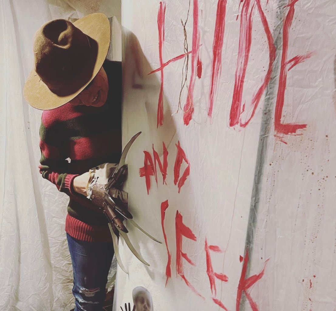 A person in a Freddy Kreuger costume next to a sheet with "Hide and Seek" written in fake blood.