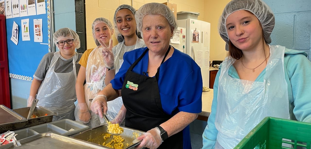 default-272 A woman in a hairnet, apron and gloves serves a scoop of macaroni and cheese, as four smiling student volunteers next to her also help distribute lunch items.