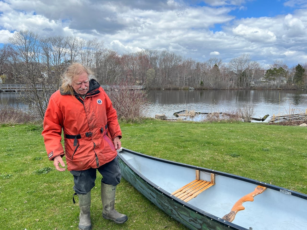 An elderly man in an orange jacket next to a small boat on the grass.