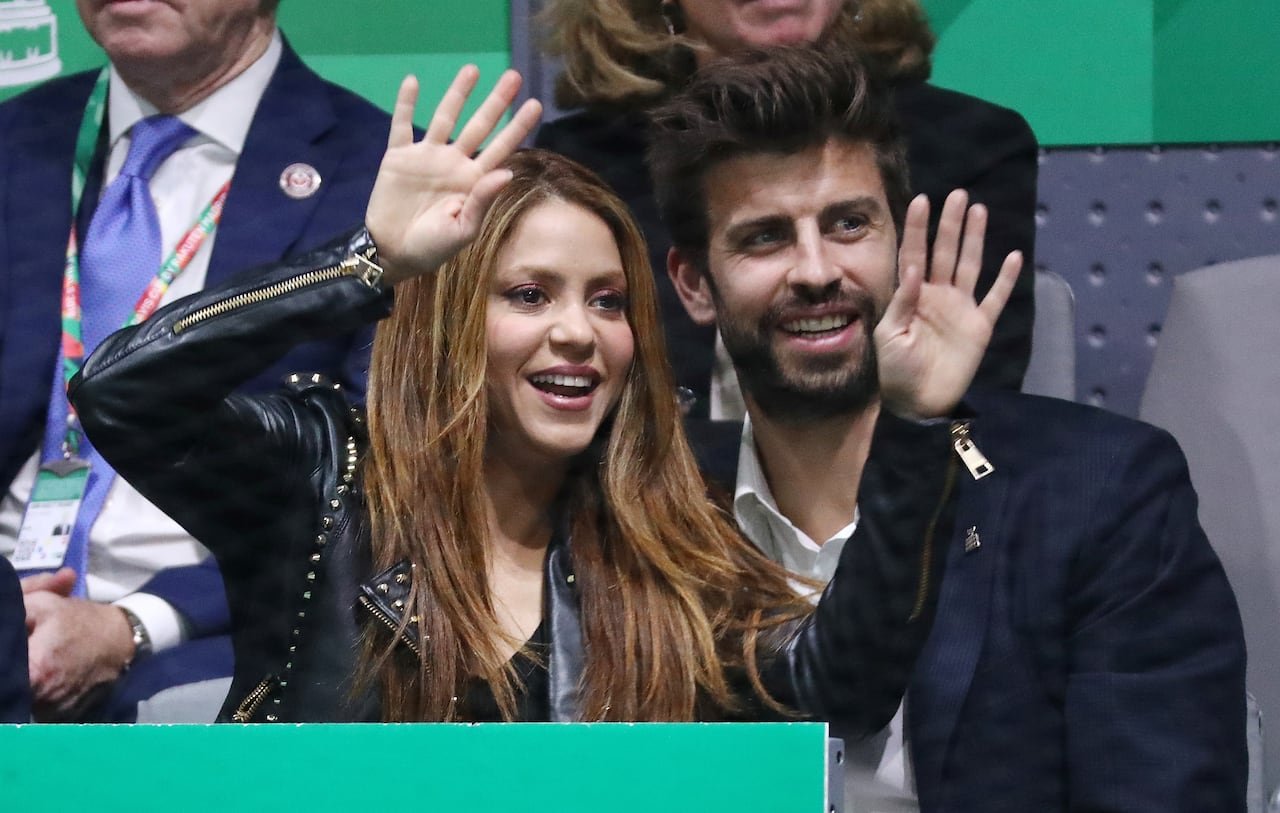 a woman with straight brown hair and a leather jackets waves with both hands from the stands at a sporting event, and a man in a dark suit jacket and white collared shirt beside her smiles