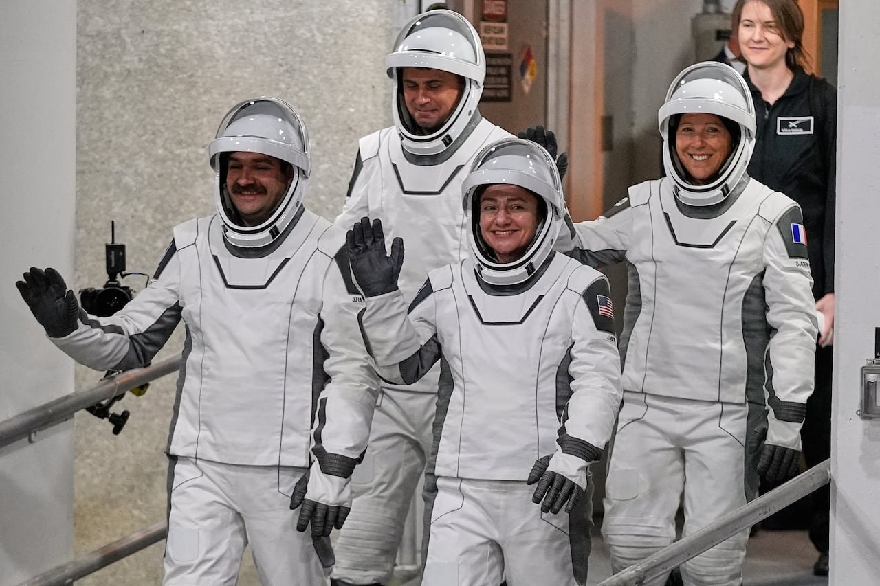 Four astronauts in their spacesuits waving and walking down a ramp.